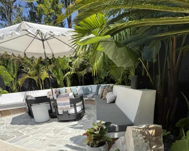The image shows a patio area with a circular stone seating arrangement, surrounded by lush greenery and an umbrella for shade.