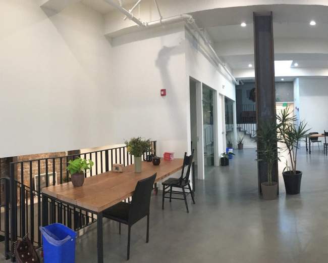 The image shows a spacious, modern interior with exposed brick walls, wooden furniture, and plants arranged in a casual office or communal space.