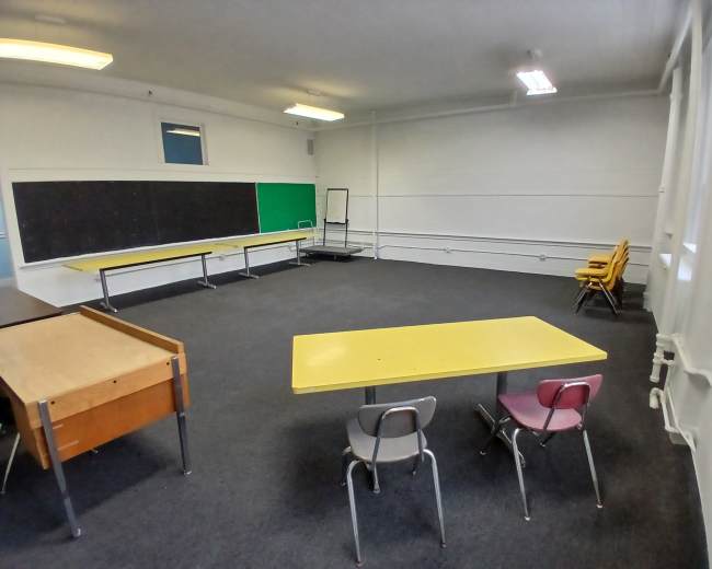 The image shows a sparsely furnished classroom with yellow and gray tables, chairs in pink and gray, and a large chalkboard on the wall.