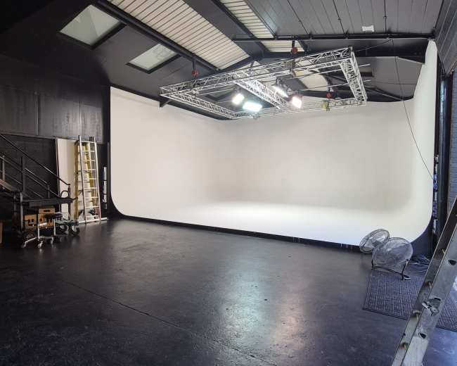 The image shows a spacious studio with a seamless white backdrop, overhead lighting rig, and equipment placed at one side.