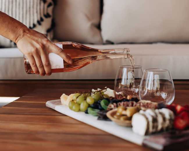 A person pours rosé wine from a bottle into two glasses next to a platter of assorted cheeses, fruits, and nuts on a wooden table.