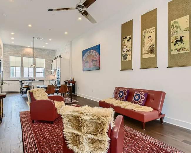 The image shows a living room with red sofas, a patterned rug, and artwork on the walls, leading into a kitchen area visible in the background.