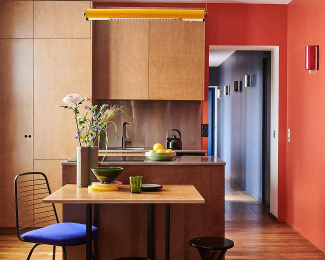 A modern kitchen features wooden cabinetry, a small dining table with black stools, and colorful accents on the walls.