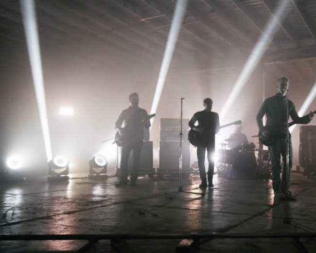 A band performs onstage in a dimly lit warehouse with bright beams of light illuminating the musicians from behind.