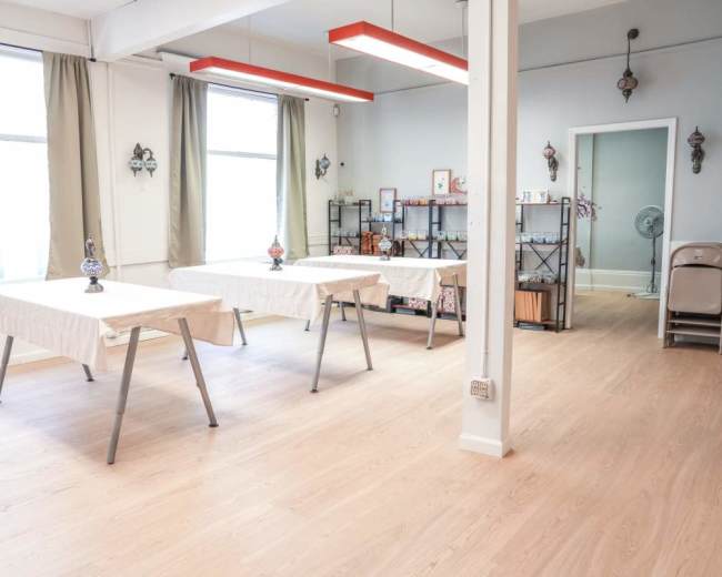 The image shows a bright, empty room with two tables covered in cloth, a decorative display shelf, and large windows letting in natural light.
