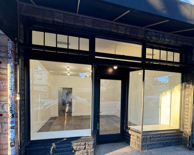 The image shows a vacant storefront with large windows and no visible signage, situated on a sidewalk.