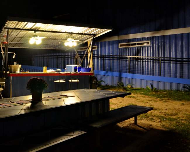 A brightly lit outdoor kitchen with a bar area and a nearby picnic table is set against a dark, wooded backdrop.