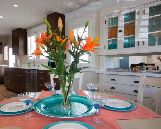 A dining table is set with plates and glasses, featuring a vase of orange lilies at the center, surrounded by a well-lit kitchen with dark cabinetry.