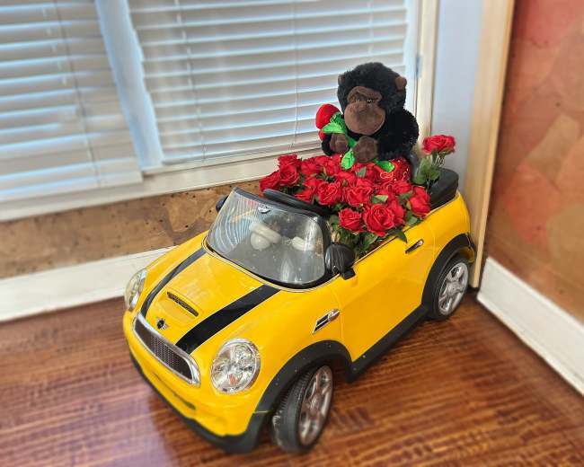A toy yellow convertible car is filled with red artificial roses and features a stuffed black gorilla sitting in the driver's seat.