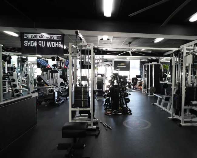 The image shows a fitness gym filled with various weight machines and exercise equipment, with signage hanging from the ceiling.