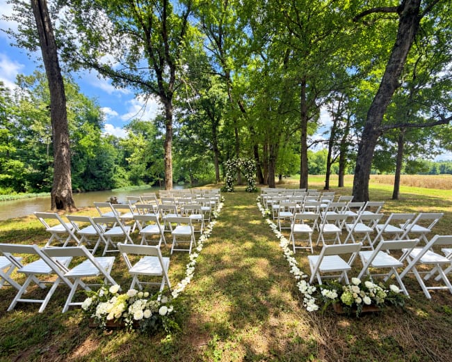 A wedding ceremony setup features rows of white chairs arranged on a grassy space under trees, with a floral arch at the end facing a serene backdrop of water and fields.