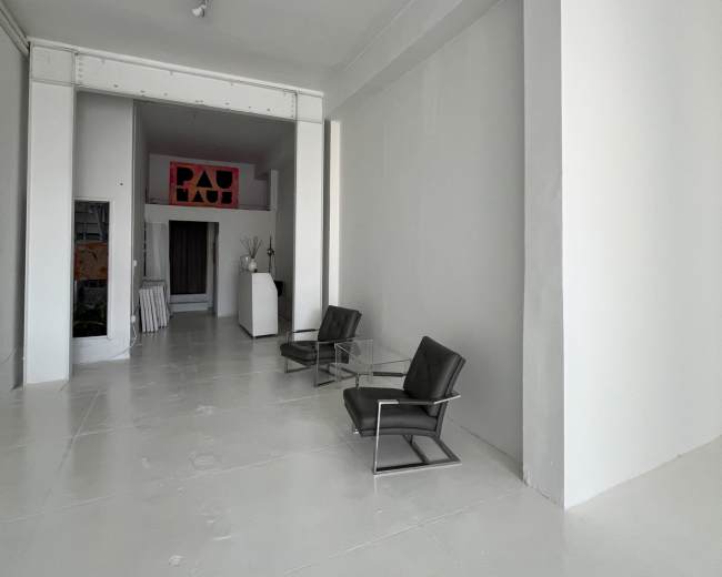 The image shows a minimalist gallery space featuring two black chairs and a glass table on a white floor, with art displayed in the background.