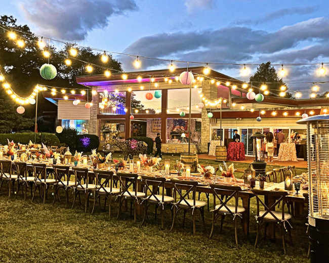 A large outdoor dining setup with a long table adorned with colorful decorations and lights, situated next to a modern building under a twilight sky.
