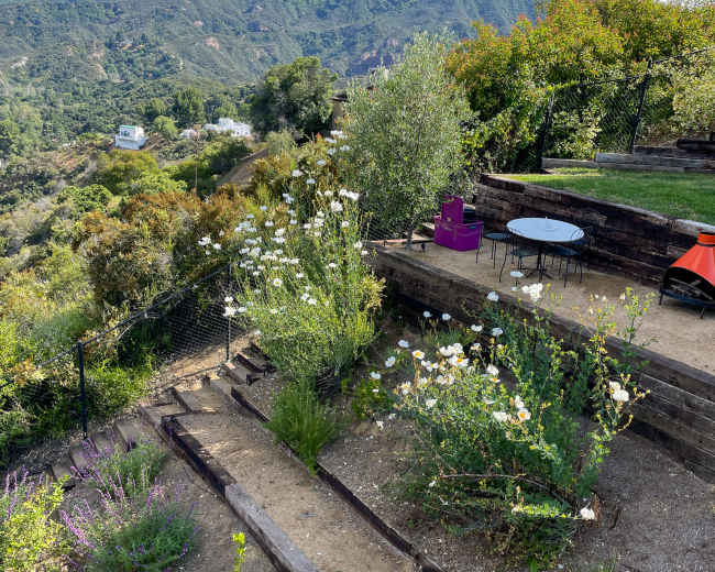 The image shows a terraced garden with flowering plants, a small table and chairs, and a backdrop of mountains under a blue sky with scattered clouds.