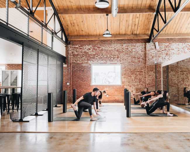 A group of individuals are participating in a workout session in a spacious studio with high ceilings and exposed brick walls.