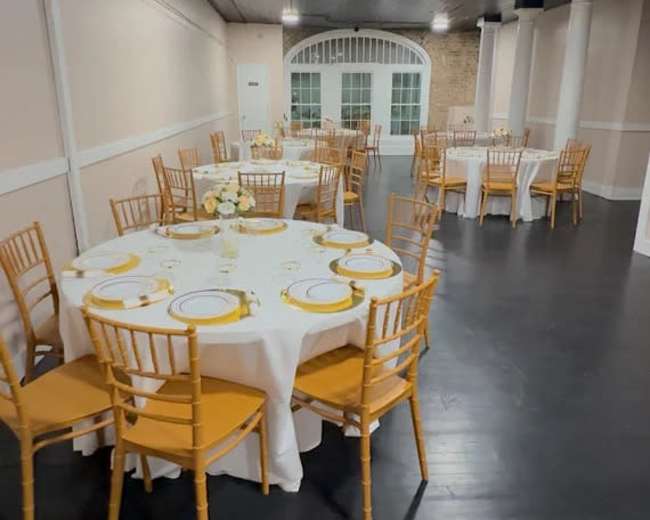 A dining area is arranged with multiple round tables, each set with white tablecloths and yellow accent chairs, in a well-lit space.