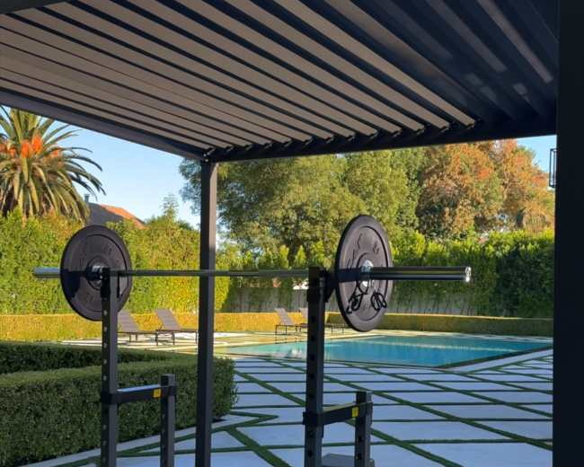 A home gym is set up under a pergola, featuring a barbell rack, workout bench, and dumbbells, overlooking a landscaped backyard with a swimming pool.