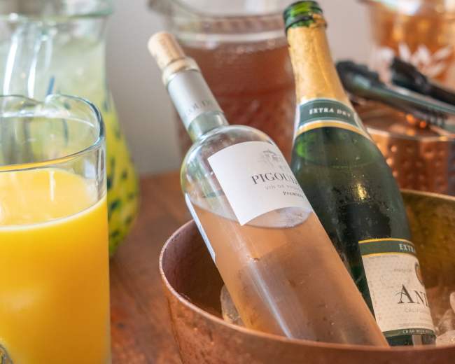A copper bowl holds two bottles of wine, one white and one sparkling, alongside a glass of orange juice and a pitcher.
