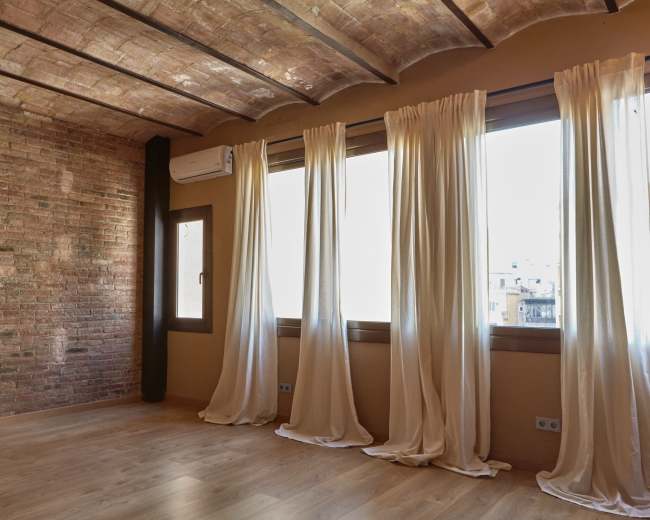 The image shows a room with exposed brick walls, large windows covered with light curtains, and wooden flooring.