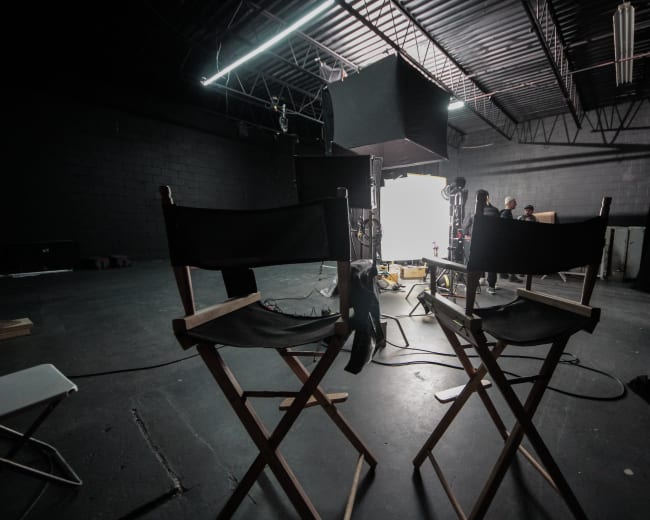 Two director chairs face a bright light source in a dimly lit studio space, with equipment and crew visible in the background.