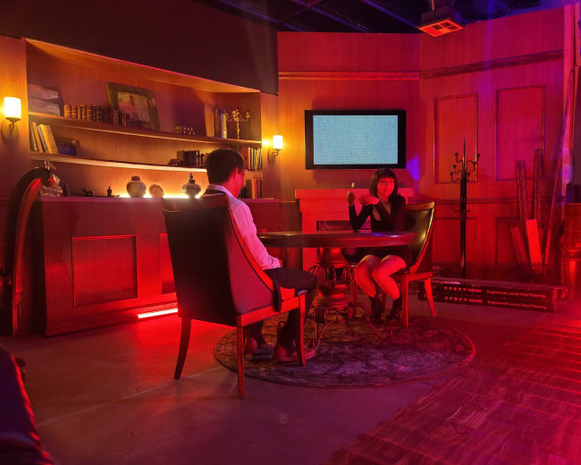A man and a woman sit across from each other at a table in a dimly lit room with red and yellow lighting, surrounded by shelves and a blank screen on the wall.
