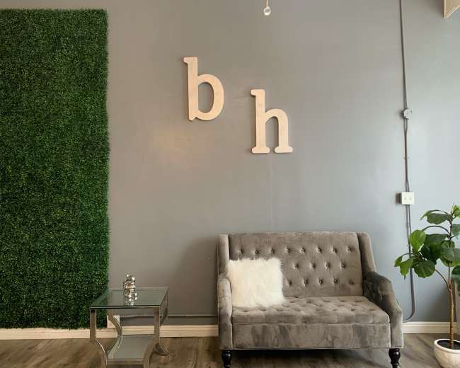 The image features a gray wall with decorative greenery on one side, a tufted gray sofa, and a glass side table with a decorative item on it.