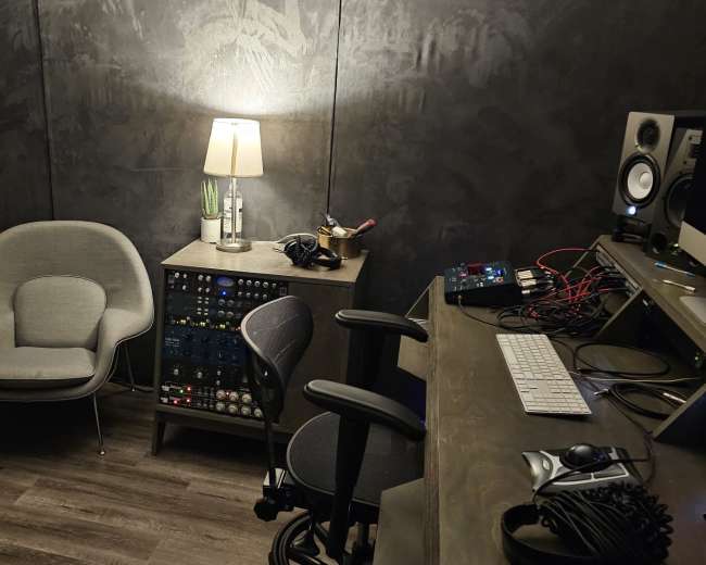 The image shows a small, dimly lit recording studio featuring two modern chairs, a lamp, a mixer console, and a computer setup against a textured wall.