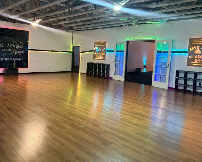 An empty dance studio features wooden flooring, colorful lights along the walls, and mirrored partitions.