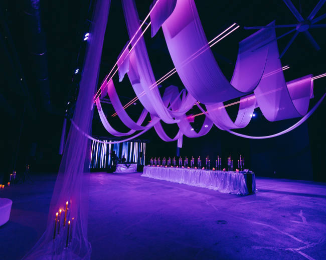 The image shows a large event space decorated with flowing fabric drapes and ambient purple lighting, featuring a long table set with candles at one end.