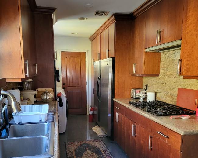 A narrow kitchen features wooden cabinets, a stainless steel refrigerator, a sink, and a long countertop with a rug running the length of the floor.