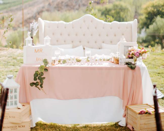 A decorated outdoor seating area features a plush, upholstered couch and a table adorned with a pink tablecloth, floral arrangements, and decorative lanterns, set against a backdrop of rolling hills and vineyards.