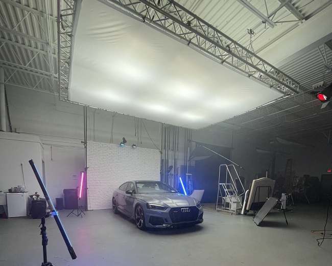 A black Audi is positioned in a studio setting with soft lighting, along with various equipment and backdrops visible.