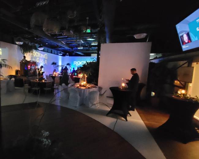 The scene shows a dimly lit event space with decorated tables, plants, and a stage area featuring colorful projections.