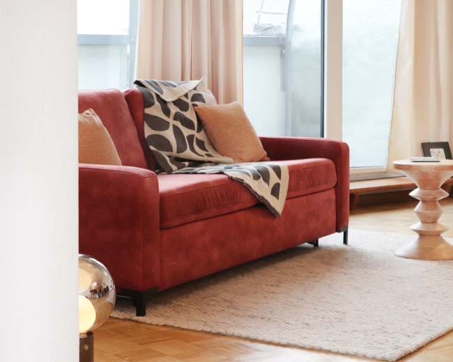 A red couch with pillows and a patterned throw is positioned in a bright living room near a textured rug and a wooden side table.