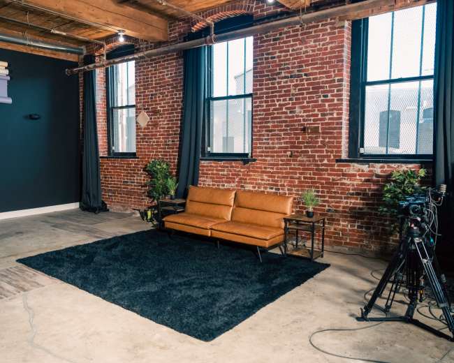 The image shows a minimalistic studio space featuring a brown leather sofa, a black rug, potted plants, and a camera setup near large windows with exposed brick walls.