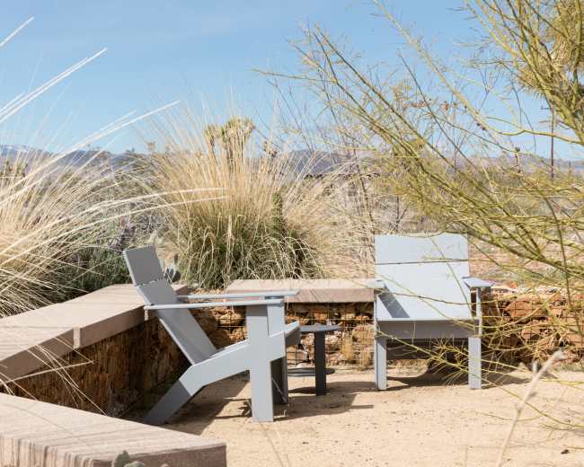 A pair of gray outdoor chairs and a small table are positioned on a stone patio surrounded by desert vegetation.