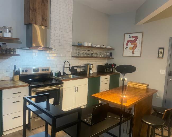 The image shows a modern kitchen with a stainless steel stove, white cabinets, and a wooden countertop, along with a small dining area featuring two stools and a laptop on the table.
