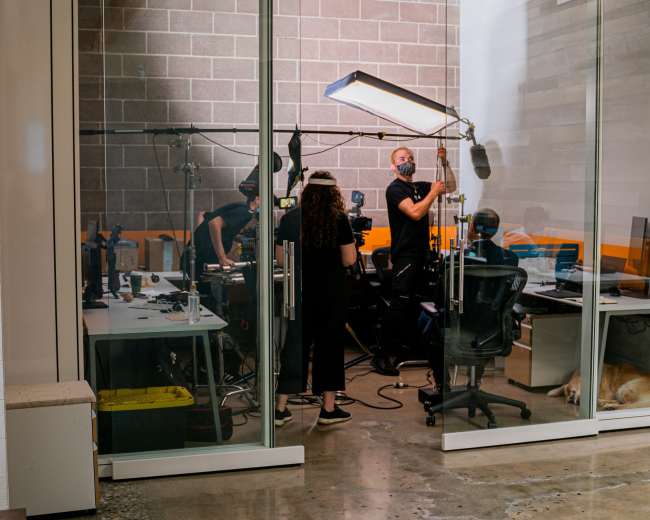 A film crew sets up equipment in a glass-enclosed office space, while a dog rests on the floor nearby.