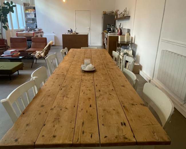 A long wooden dining table with white chairs is set in a well-lit room with minimal decor and exposed light bulbs hanging from the ceiling.