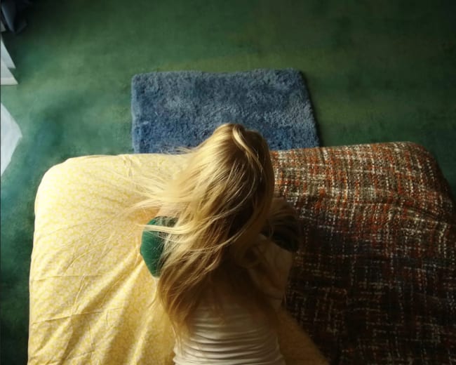 A person with long hair is sitting on a bed, facing away, with a blanket partially covering them.