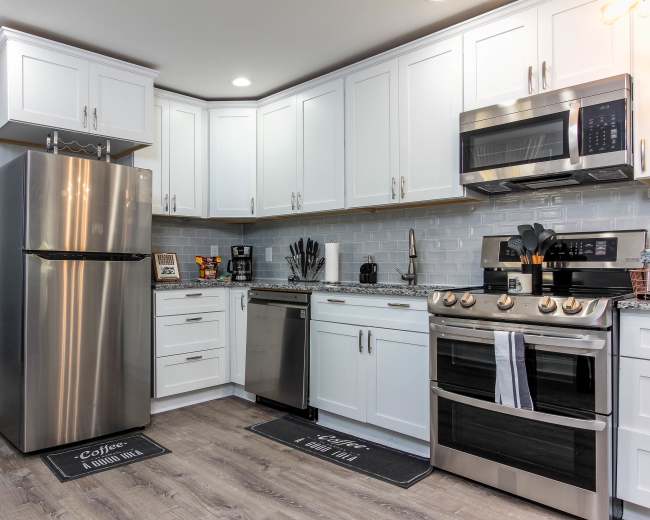 The image shows a modern kitchen featuring white cabinetry, stainless steel appliances, and a gray tiled backsplash.