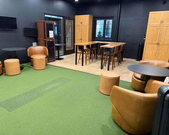 The image shows a lounge area with brown furniture, including circular and rectangular tables, seating options, and green carpet, located in a room with black walls.