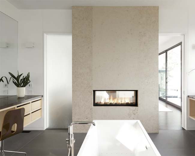 A modern bathroom features a freestanding bathtub, a minimalist fireplace embedded in a textured wall, and large windows allowing natural light.