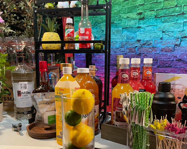 A variety of beverage ingredients and mixers are arranged on a bar cart beside a cutting board with a knife and a glass filled with lemons and limes.