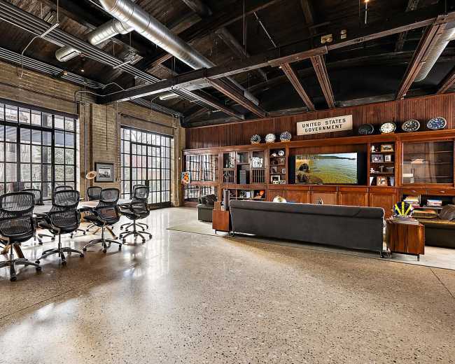 The image shows a spacious industrial-style office with large windows, a conference table with chairs, and a wooden wall featuring shelves of decor and artifacts.