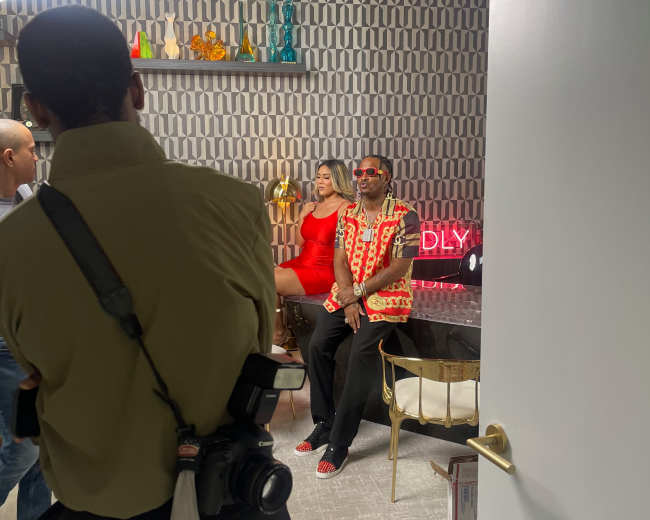 A photoshoot is taking place in a stylized room with a man in sunglasses and a patterned shirt sitting next to a woman in a red dress, while a photographer captures the scene.