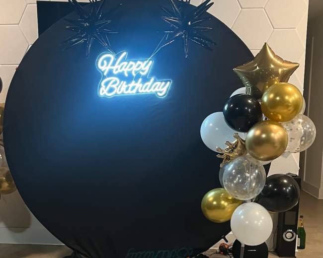 A black circular backdrop featuring a neon "Happy Birthday" sign is decorated with gold and black balloons on one side.