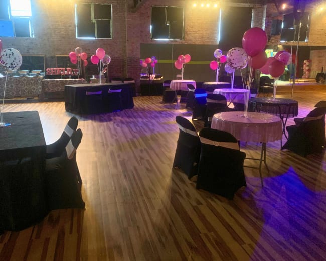 A decorated event space with tables and chairs, featuring balloons and a wooden floor.