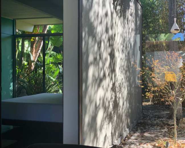 The image shows a view from inside a modern structure, featuring a bed and a bright window that looks out onto a garden with tropical plants.