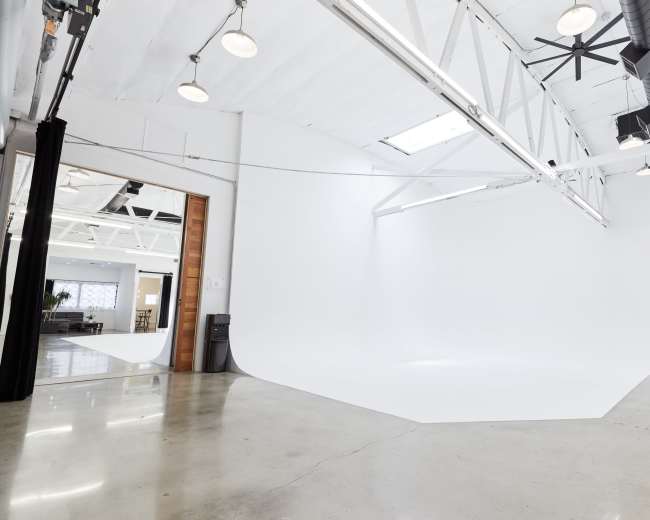 The image depicts a large, empty studio space with a seamless white backdrop, polished concrete floors, and industrial lighting.